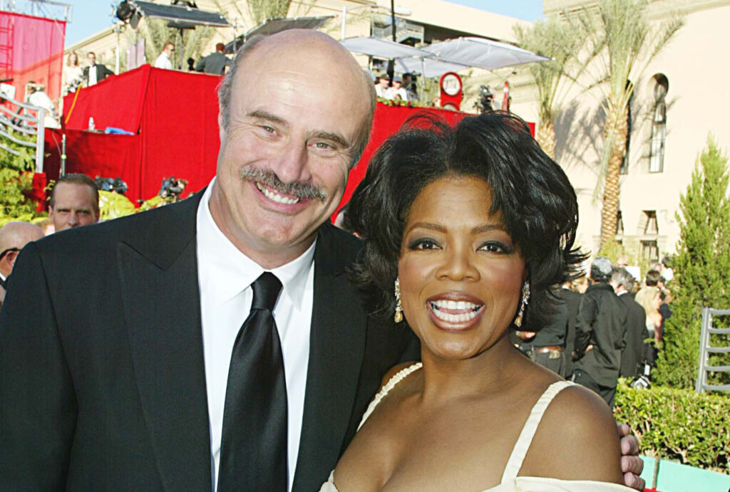Phil McGraw, in a black suit and black tie, stands with Oprah Winfrey, in a yellow dress and drop earrings, as both smile on a red carpet with red staging, palm trees and photographers behind them.