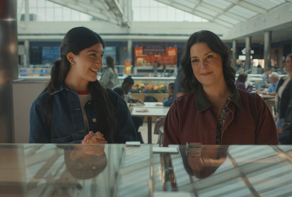 Two women stand together at a jewelry counter in a mall food court, one in a denim jacket smiling and the other in a maroon jacket looking on, in a scene from